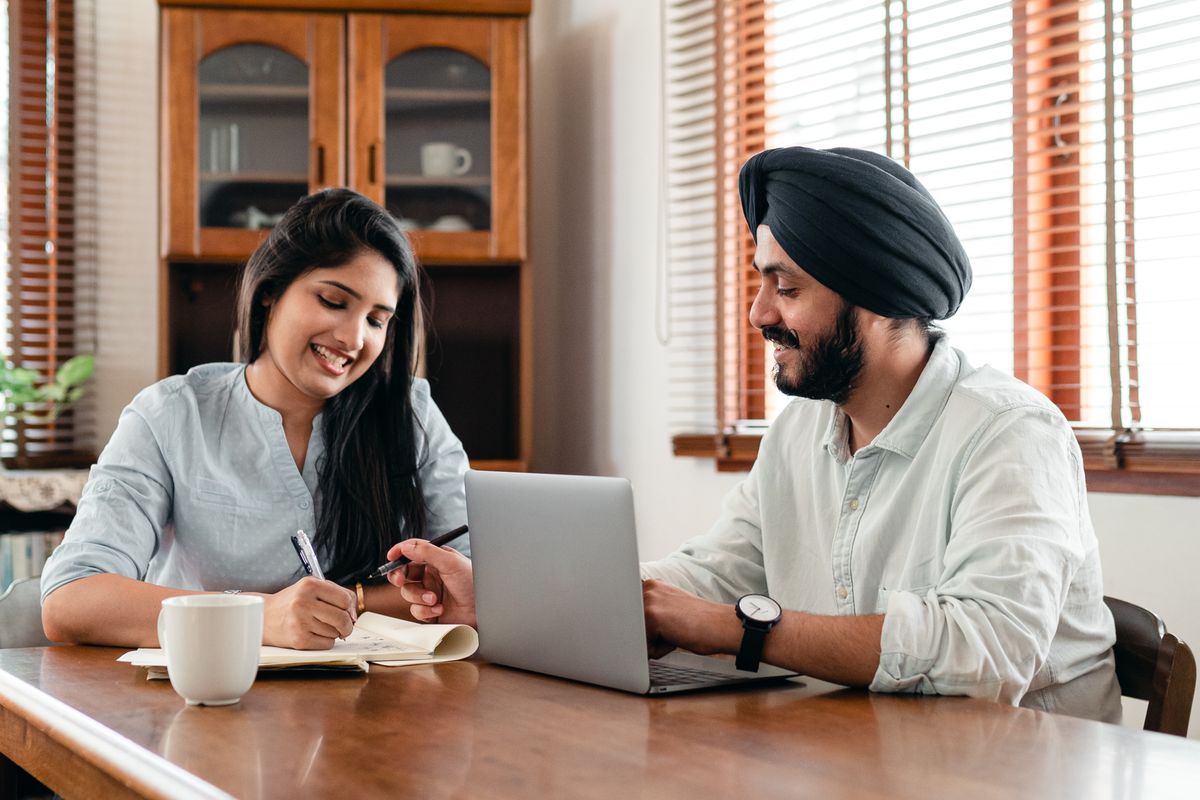 Two colleagues, a woman and a man in a turban, working together over a laptop and notebook